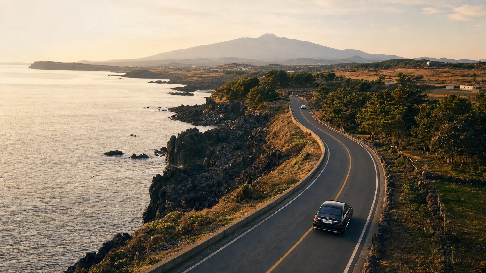 Black Mercedes-Benz S-Class sedan on Jeju's volcanic coastal road with Hallasan in the distance at golden hour